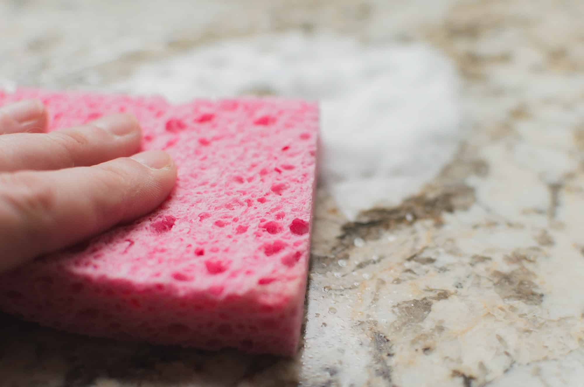 Hand using a pink sponge to wipe a granite counter with foaming cleanser, doing household chores